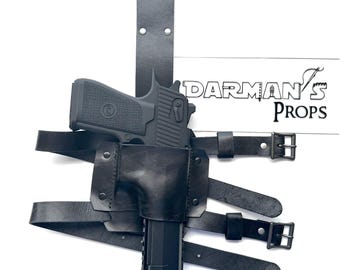 May include: Black leather holster with a black handgun and a black strap. The holster is designed to be worn on the thigh. The strap is adjustable and has a buckle. The holster is labeled "Darman's Props".