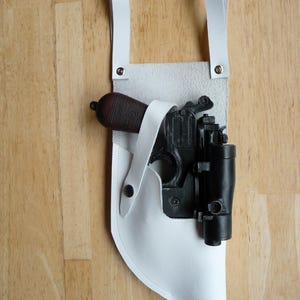 May include: White leather holster with a black and brown toy gun. The holster has two straps and a snap closure.