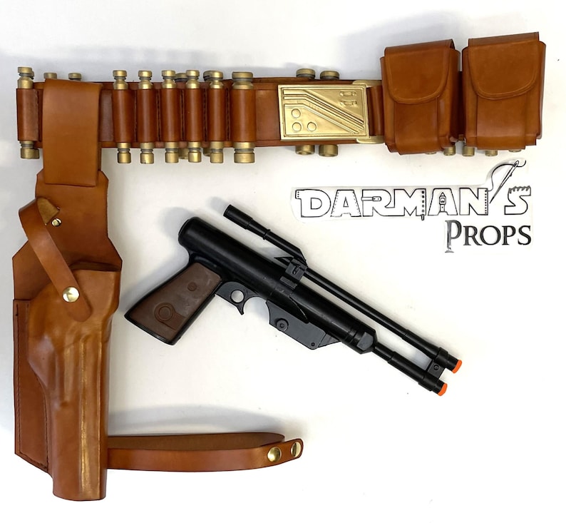 May include: A brown leather belt with a gold buckle and a holster for a black pistol. The belt has a row of brass cartridges attached to it. There are two additional brown leather pouches attached to the belt. The holster is attached to a brown leather strap. The text "DARMAN'S PROPS" is visible in the image.