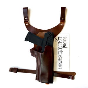 Puede incluir: Un holster de hombro de cuero marrón con una pistola negra en su interior. El holster tiene una correa que se coloca sobre el hombro y una hebilla en el lateral. El holster está etiquetado como "DARMAN'S PROPS".