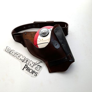 May include: A brown leather holster with a black strap and a silver buckle. The holster is designed to hold a round, white and red object with a black and silver design. The holster is attached to a brown leather belt with silver rivets.