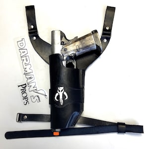 May include: A black leather holster with a silver pistol inside. The holster features a Mandalorian symbol on the front and is attached to a black leather shoulder strap. The strap has a buckle and a D-ring. The holster is labelled "Darman's Props".