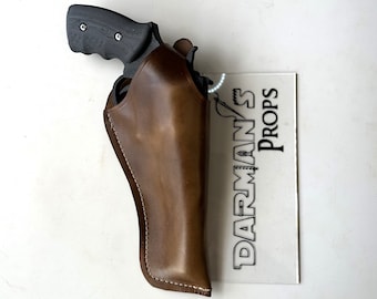 May include: A brown leather holster with a black handgun inside. The holster is stitched with white thread and has a flap that covers the gun. The holster is on a clear acrylic surface with the text "DARMAN'S PROPS" printed on it.