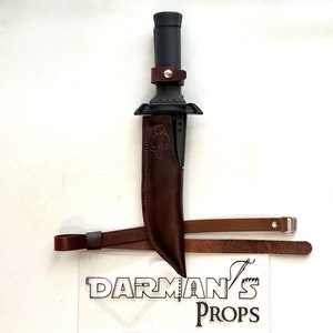 Puede incluir: Un cuchillo negro con una funda de cuero marrón y una correa de cuero marrón. El cuchillo tiene un mango negro y un anillo de metal plateado. La correa tiene una hebilla de metal plateado. El cuchillo está etiquetado como "DARMAN'S PROPS".