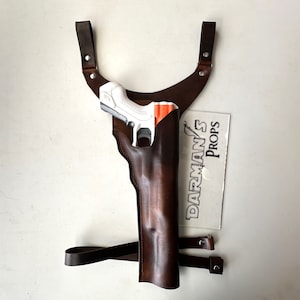 Könnte beinhalten: Ein braunes Schulterholster aus Leder mit einer weißen und orangefarbenen Spielzeugpistole. Das Holster hat verstellbare Riemen und ein vertikales Design. Das Bild enthält auch ein klares Schild mit dem Text "DARMAN'S PROPS".