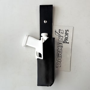May include: A white plastic toy gun in a black leather holster with a strap. The holster is hanging on a white wall. A business card with the text "DARMAN'S PROPS" is visible in the background.