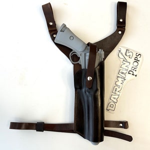 May include: A brown leather holster with a gray gun inside. The holster has a strap that goes over the shoulder and a belt strap. The holster is labeled "DARMAN'S PROPS".