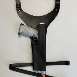 Könnte beinhalten: Ein schwarzes Leder-Schulterholster mit einer silbernen Pistole darin. Das Holster hat einen Riemen, der um die Taille geht, und einen Riemen, der über die Schulter geht.