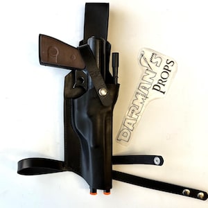 May include: A black leather holster with a gun and a strap. The holster has a metal buckle and a small, clear plastic tag with the text "DARMAN'S PROPS" printed on it.