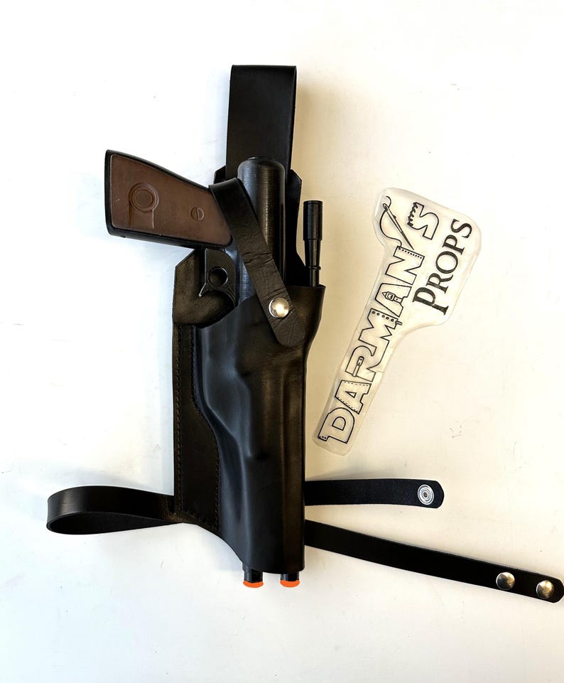 May include: A black leather holster with a gun and a strap. The holster has a metal buckle and a small, clear plastic tag with the text "DARMAN'S PROPS" printed on it.