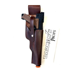 May include: A dark brown leather holster containing a black and tan prop blaster. The holster features a vertical strap with a silver metal snap button. Next to the holster is a white card with the black text DARMAN'S PROPS printed vertically.