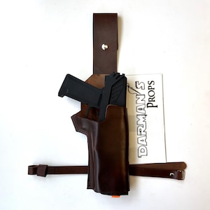 Puede incluir: Una funda de cuero marrón con una pistola negra en su interior. La funda tiene una correa que se ajusta a la cintura. La funda está etiquetada como "Darman's Props".