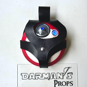 Op de afbeelding: Een zwart leren rekwisiet met een rood en wit cirkelvormig ontwerp, met een lens en een blauw licht. De rekwisiet wordt op zijn plaats gehouden door een zwarte leren riem en een zilveren knoop. De tekst "DARMAN'S PROPS" is zichtbaar.