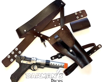 May include: A brown leather holster with a silver buckle and a black gun with a silver barrel. The holster is attached to a brown leather belt with silver studs. The belt is also attached to a brown leather strap with a silver buckle. The strap is worn over the shoulder. The text "DARMAN'S PROPS" is visible in the image.