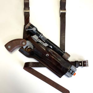 Op de afbeelding: Een bruine leren holster met een pistool en een richtkijker. De holster heeft twee banden die over de schouder gedragen kunnen worden. De banden zijn van bruin leer en hebben metalen gespen.