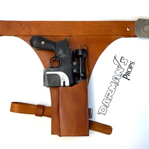 May include: A brown leather holster with a black and silver blaster pistol and a scope attached. The holster has a strap and buckle. The text "DARMAN'S PROPS" is visible in the lower right corner of the image.