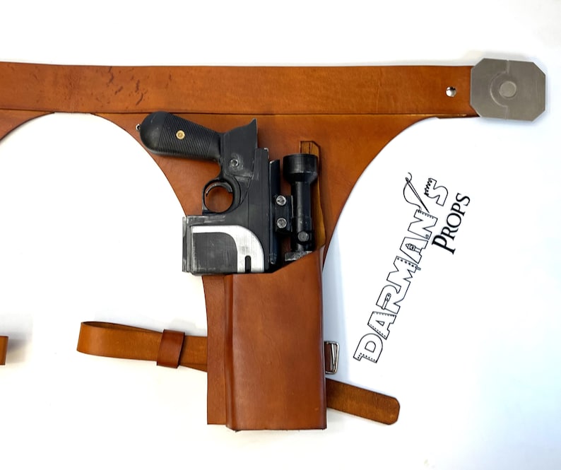 May include: A brown leather holster with a black and silver blaster pistol and a scope attached. The holster has a strap and buckle. The text "DARMAN'S PROPS" is visible in the lower right corner of the image.