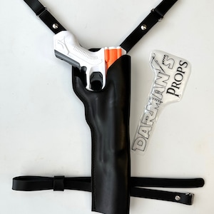 Könnte beinhalten: Ein schwarzes Lederholster mit einer weißen und orangefarbenen Spielzeugpistole darin. Das Holster hat einen schwarzen Riemen, der über die Schulter geht. Der Riemen hat silberne Metallnieten. Ein weißer Aufkleber mit schwarzem Text, der "DARMAN'S PROPS" lautet, befindet sich auf der rechten Seite des Bildes.