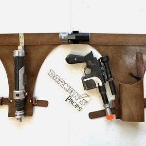 May include: A brown leather belt with two holsters. One holster holds a silver and black lightsaber, and the other holds a black and silver blaster pistol. The belt has a metal buckle and a metal loop on the right side. The belt is labeled "Darman's Props".