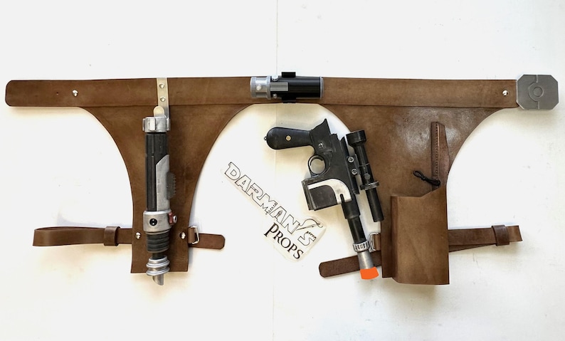 May include: A brown leather belt with two holsters. One holster holds a silver and black lightsaber, and the other holds a black and silver blaster pistol. The belt has a metal buckle and a metal loop on the right side. The belt is labeled "Darman's Props".