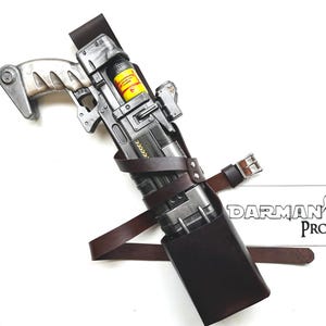 May include: A prop gun with a silver and black finish, featuring a brown leather strap and a holster. A yellow and black container is visible. The handle has a unique, textured design. The text "DARMAN'S PROPS" is in the lower right corner.