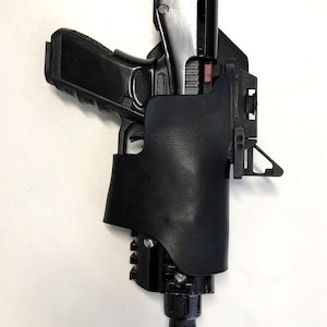 May include: A black leather holster with a black pistol inside. The holster has a metal clip and a black plastic base.