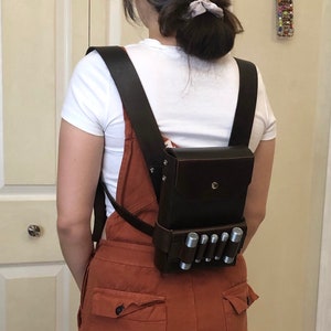 Może przedstawiać: Brązowy skórzany plecak z dużą kieszenią z przodu i rzędem srebrnych cylindrów. Plecak nosi osoba ubrana w białą koszulę i brązowe ogrodniczki.