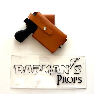 May include: A brown leather holster with a black toy gun inside. The holster has a silver snap button and a strap. The image also includes a clear acrylic sign with the text "DARMAN'S PROPS" in black.