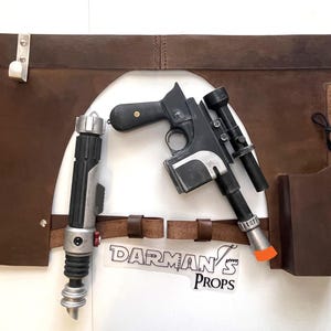 May include: A brown leather belt with two holsters, one for a blaster pistol and one for a lightsaber. The belt is decorated with metal rivets and has a metal buckle. The blaster pistol is black and silver with a scope. The lightsaber is black and silver with a red blade.