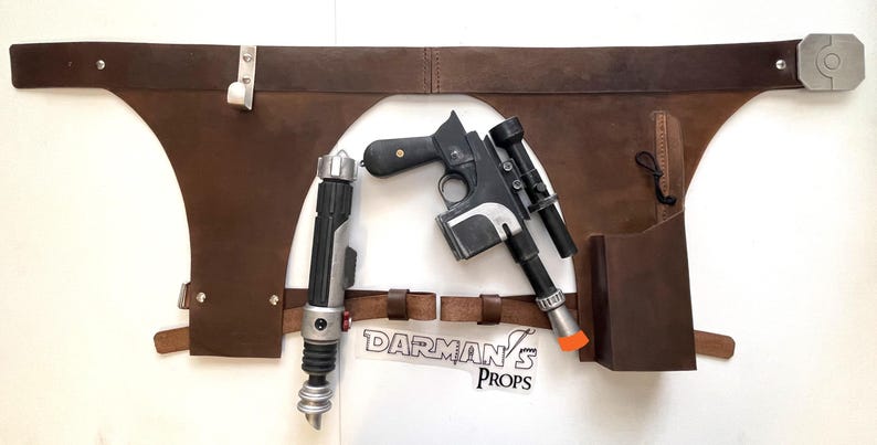 May include: A brown leather belt with two holsters, one for a blaster pistol and one for a lightsaber. The belt is decorated with metal rivets and has a metal buckle. The blaster pistol is black and silver with a scope. The lightsaber is black and silver with a red blade.