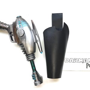May include: A silver and gray ray gun prop with blue accents and a black holster. The ray gun has a detailed design with a ribbed handle, a trigger, and a fin-like structure. The holster is made of black material.