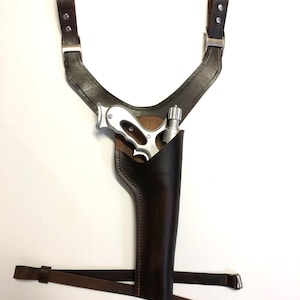 May include: A brown leather holster with a silver gun inside. The holster is attached to a brown leather shoulder strap.