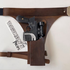 May include: A brown leather holster with a black strap and a brass buckle. A black and silver blaster pistol is in the holster. The holster is labeled "Darman's Props".