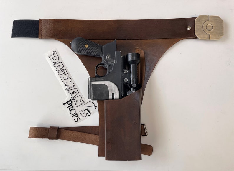 May include: A brown leather holster with a black strap and a brass buckle. A black and silver blaster pistol is in the holster. The holster is labeled "Darman's Props".