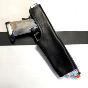以下が含まれることがあります： 黒い革製のホルスターの中に、銀色と茶色の拳銃が入っています。ホルスターは黒い革製のベルトに取り付けられています。