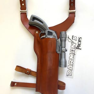May include: A brown leather holster with a silver gun and a scope attached. The holster has a strap that goes over the shoulder. The text "DARMAN'S Props" is visible on a white sticker.