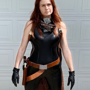 May include: A woman wearing a black leather jumpsuit with a brown leather belt and a brown leather holster. She is also wearing black leather gloves and goggles.