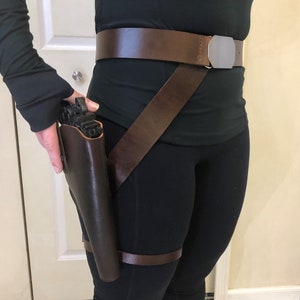 May include: A brown leather belt with a silver buckle and a matching thigh holster. The holster is worn on the left leg and has a strap that goes over the right shoulder.