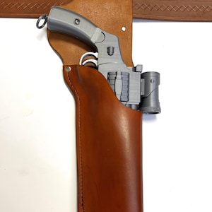 May include: A gray revolver in a brown leather holster attached to a brown belt. The holster is stitched and riveted, with a loop for the gun's handle. The belt has a decorative pattern.