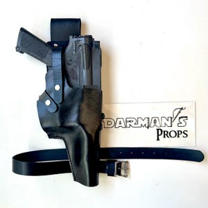 May include: Black leather holster with a gun inside, secured by a belt. The holster has a molded design and is held together with metal rivets. The belt is black with a silver buckle. The text "DARMAN'S PROPS" is visible.