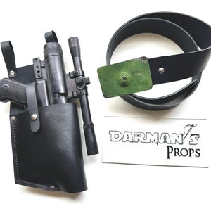 May include: A black leather holster with a blaster and scope, and a black belt with a green buckle. The holster has a strap and is attached to a belt. The text "DARMAN'S PROPS" is visible.