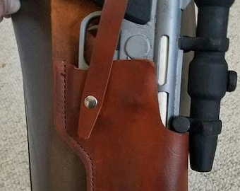 May include: A brown leather holster with a silver and black pistol and a black scope attached. The holster has a strap and a metal button.