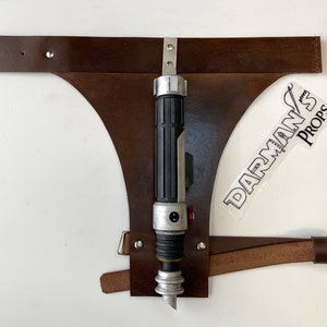 May include: A brown leather belt with a metal buckle and a silver and black lightsaber attached to it. The belt is labeled "DARMAN'S PROPS".