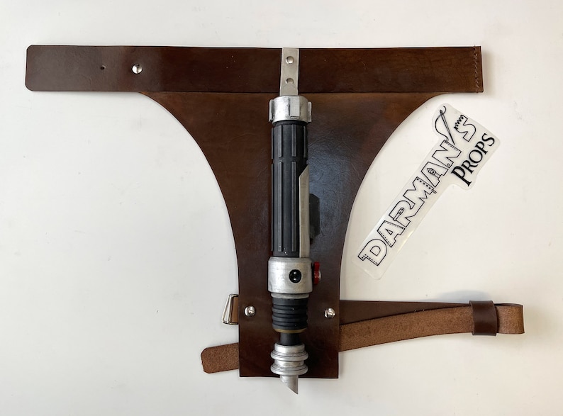May include: A brown leather belt with a metal buckle and a silver and black lightsaber attached to it. The belt is labeled "DARMAN'S PROPS".