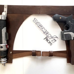 May include: A brown leather belt with two holsters. The left holster holds a black and silver lightsaber. The right holster holds a black blaster pistol with a scope.
