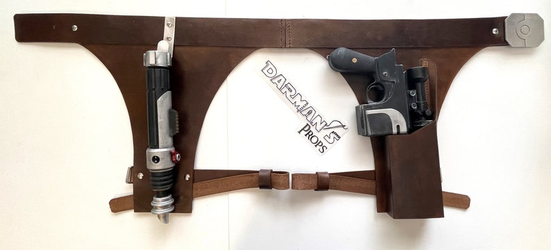 May include: A brown leather belt with two holsters. The left holster holds a black and silver lightsaber. The right holster holds a black blaster pistol with a scope.