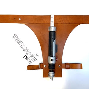 May include: A brown leather belt with a holster for a lightsaber. The holster is attached to the belt with metal rivets. The lightsaber is silver and black with a red button.