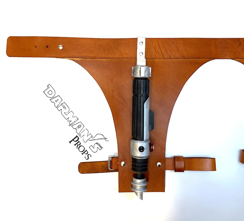 May include: A brown leather belt with a holster for a lightsaber. The holster is attached to the belt with metal rivets. The lightsaber is silver and black with a red button.
