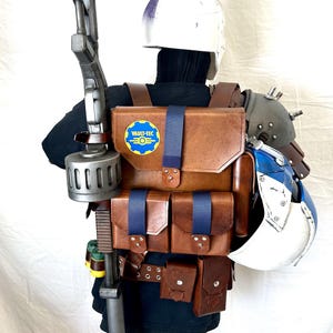 May include: A brown leather backpack with a blue strap and a Vault-Tec logo patch. The backpack has multiple pockets and is worn by a figure wearing a white helmet and a blue and white armoured suit.