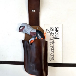 May include: A brown leather holster with a strap for a Nerf gun. The holster is hanging on a white wall. The Nerf gun is in the holster. The text "DARMAN'S PROPS" is visible on a white card.